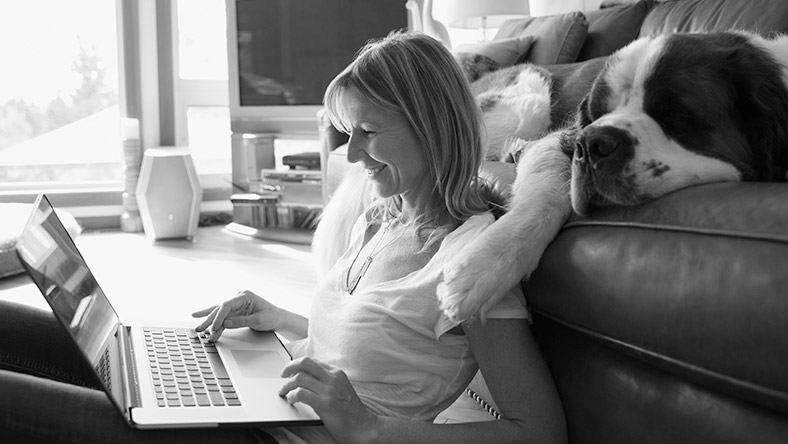 Woman sitting the floor, with a laptop on her lap and a dog laying behind her.