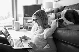 Woman sitting the floor, with a laptop on her lap and a dog laying behind her.