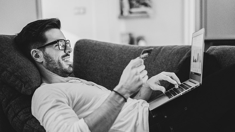 Man wearing glasses laying on a couch with his credit card and laptop.