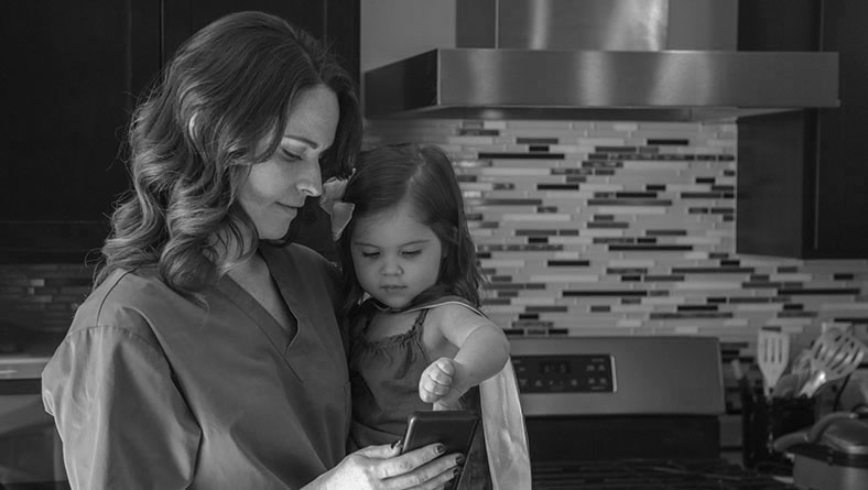 A nurse holding her daughter standing in a kitchen looking at a mobile phone.