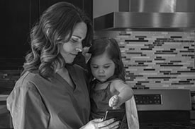 A nurse holding her daughter standing in a kitchen looking at a mobile phone.